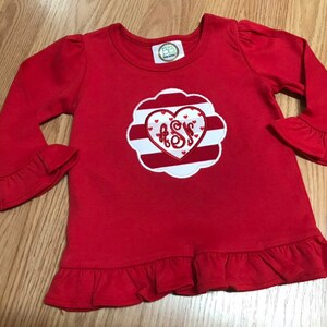K&ouml;nnte beinhalten: Rotes Langarm-Shirt mit einem wei&szlig;-rot gestreiften Herzen, in dessen Mitte die Buchstaben "ASS" gestickt sind. Das Shirt hat R&uuml;schen an den &Auml;rmeln und am Saum.