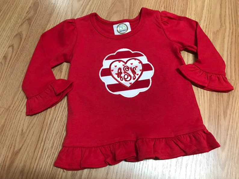 K&ouml;nnte beinhalten: Rotes Langarm-Shirt mit einem wei&szlig;-rot gestreiften Herzen, in dessen Mitte die Buchstaben "ASS" gestickt sind. Das Shirt hat R&uuml;schen an den &Auml;rmeln und am Saum.