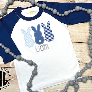 May include: White baseball tee with blue raglan sleeves, featuring three blue and white bunny appliques and the name "Liam" embroidered on the front. The shirt has a "Made with Love Marketplace" logo on the bottom.