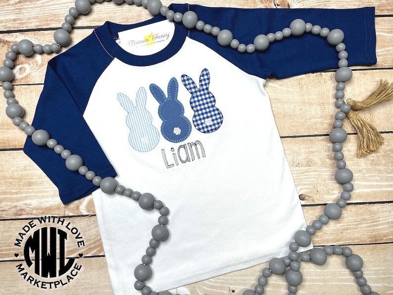 May include: White baseball tee with blue raglan sleeves, featuring three blue and white bunny appliques and the name "Liam" embroidered on the front. The shirt has a "Made with Love Marketplace" logo on the bottom.