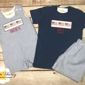 May include: Two sets of matching baby clothes with a blue and white striped pattern. One set includes a sleeveless romper with the name "BASTON" and a row of American flags embroidered on the front. The other set includes a navy blue t-shirt with the name "ELI" and a row of American flags embroidered on the front, and matching blue and white striped shorts.