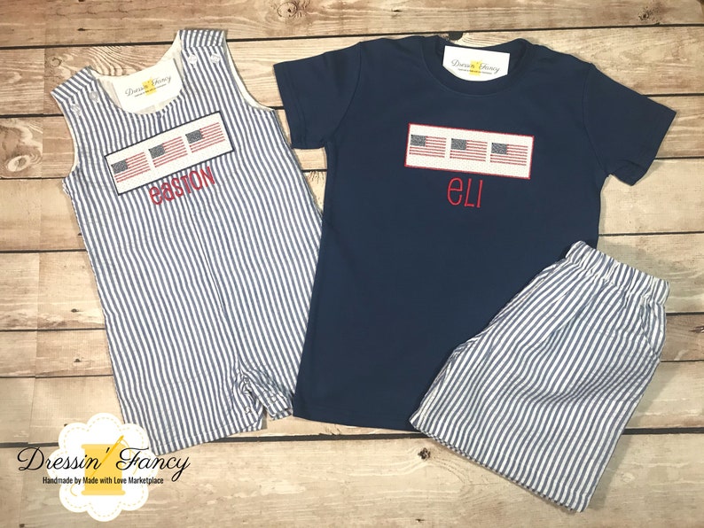 May include: Two sets of matching baby clothes with a blue and white striped pattern. One set includes a sleeveless romper with the name "BASTON" and a row of American flags embroidered on the front. The other set includes a navy blue t-shirt with the name "ELI" and a row of American flags embroidered on the front, and matching blue and white striped shorts.
