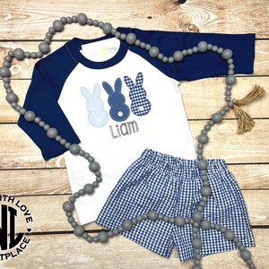 May include: A white and navy blue baseball tee shirt with three blue bunny appliques and the name "Liam" embroidered on the front. The shirt is paired with blue and white gingham shorts. A grey beaded necklace with tassels is included.