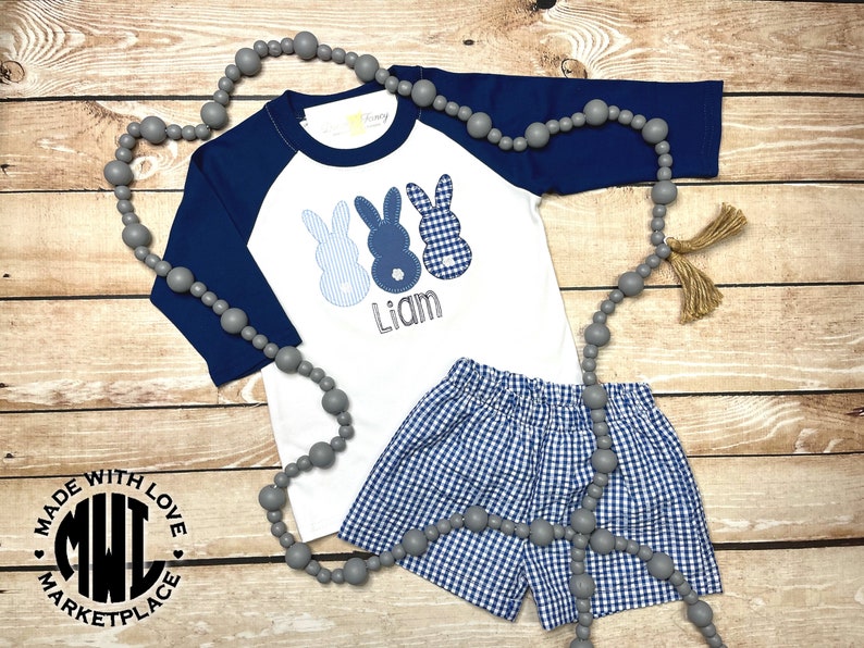 May include: A white and navy blue baseball tee shirt with three blue bunny appliques and the name "Liam" embroidered on the front. The shirt is paired with blue and white gingham shorts. A grey beaded necklace with tassels is included.