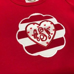K&ouml;nnte beinhalten: Rot-wei&szlig; gestreiftes Herz mit einem wei&szlig;en Monogramm "A.S.M." im Inneren. Das Herz ist von einem wei&szlig;en, gewellten Rand umgeben.
