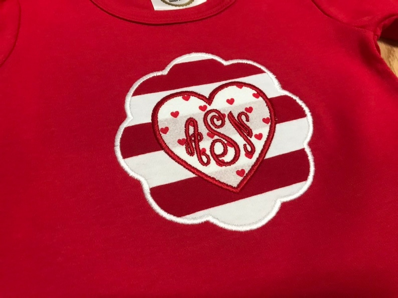K&ouml;nnte beinhalten: Rot-wei&szlig; gestreiftes Herz mit einem wei&szlig;en Monogramm "A.S.M." im Inneren. Das Herz ist von einem wei&szlig;en, gewellten Rand umgeben.