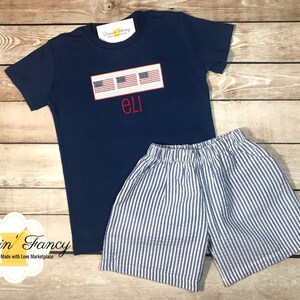 Pu&ograve; includere: Una maglietta blu navy con una fila di quattro piccole bandiere americane e il nome "ELI" ricamato in rosso. La maglietta &egrave; abbinata a pantaloncini a righe blu e bianche.