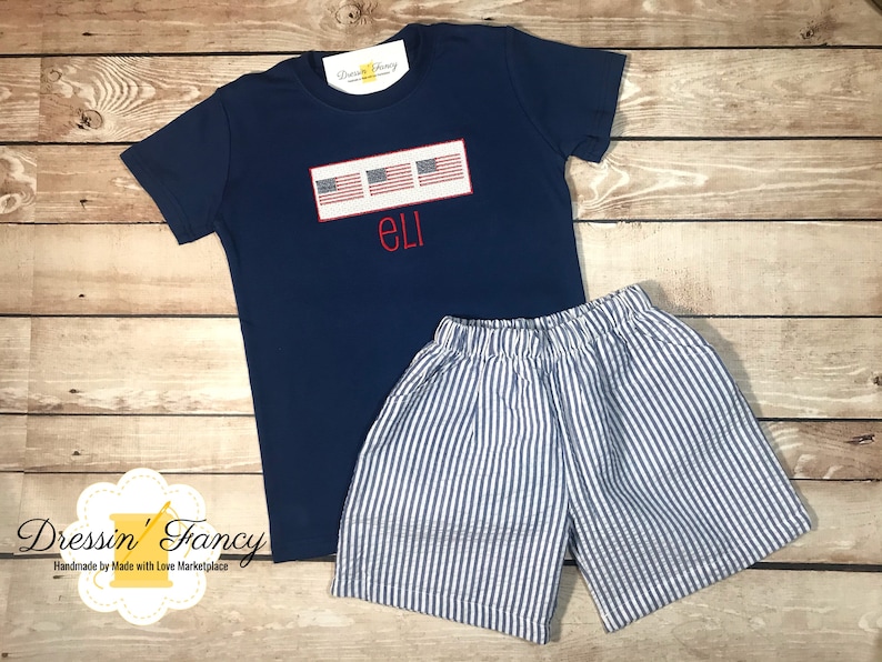 Pu&ograve; includere: Una maglietta blu navy con una fila di quattro piccole bandiere americane e il nome "ELI" ricamato in rosso. La maglietta &egrave; abbinata a pantaloncini a righe blu e bianche.