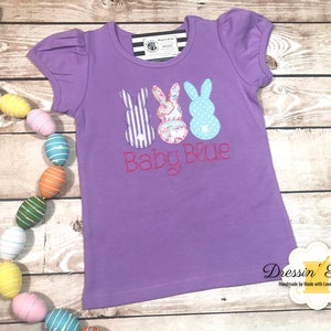 Puede incluir: Camiseta morada de manga corta con tres aplicaciones de conejito y el texto "Baby Blue" en rosa.