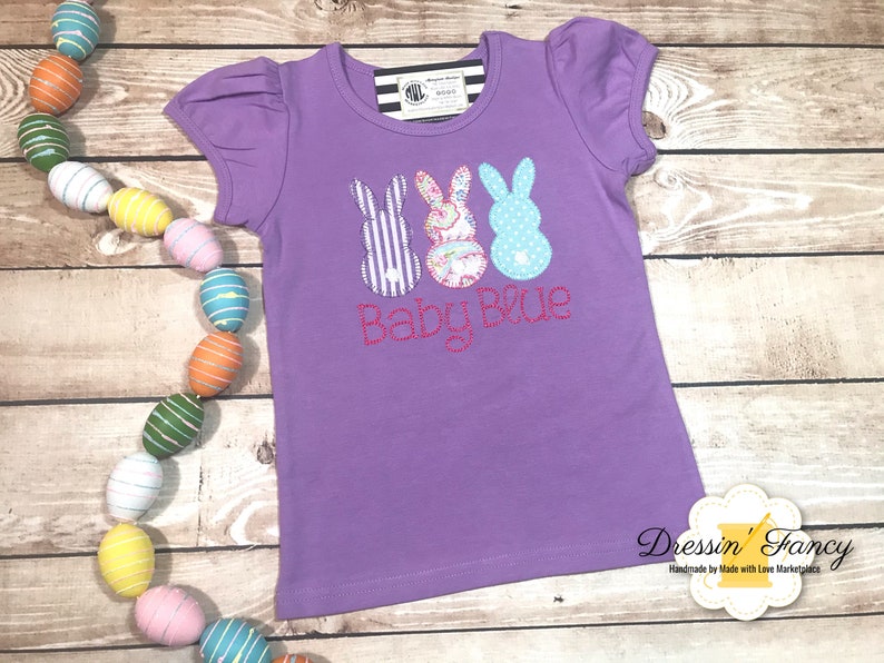 Puede incluir: Camiseta morada de manga corta con tres aplicaciones de conejito y el texto "Baby Blue" en rosa.