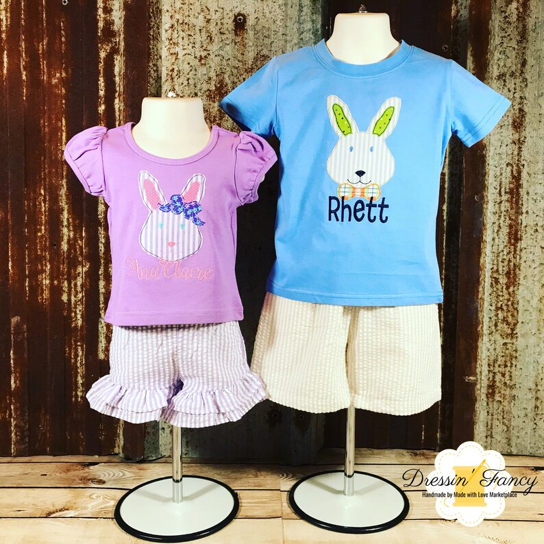May include: Two children's outfits with embroidered bunny designs. The girl's outfit is purple with a pink bow on the bunny. The boy's outfit is blue with a bow tie on the bunny. Both outfits have the names "Anna Claire" and "Rhett" embroidered on the shirts.