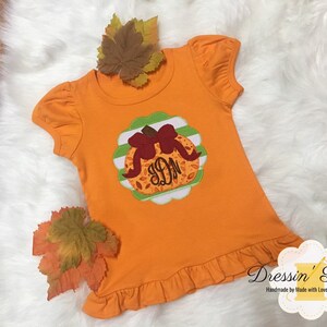 May include: Orange short-sleeved shirt with a pumpkin applique featuring a brown bow and initials. The applique is set on a scalloped green and white background. The shirt has a ruffled hem and sleeves. Decorative fall leaves are placed around the shirt.