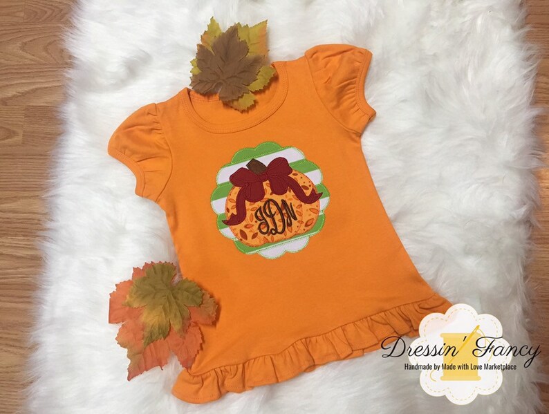 May include: Orange short-sleeved shirt with a pumpkin applique featuring a brown bow and initials. The applique is set on a scalloped green and white background. The shirt has a ruffled hem and sleeves. Decorative fall leaves are placed around the shirt.