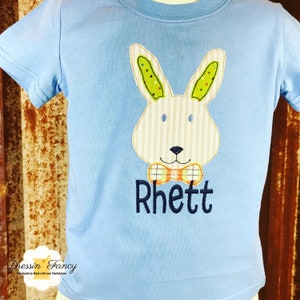 May include: A light blue t-shirt with a white bunny applique with green ears and a plaid bow tie. The name "Rhett" is embroidered in navy blue below the bunny.