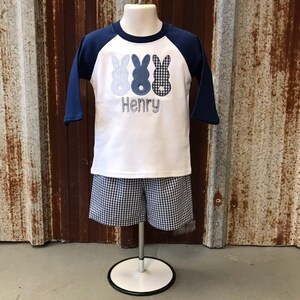 May include: A white and blue baseball style shirt with three blue bunny silhouettes and the name "Henry" embroidered on the front. The shirt is paired with blue and white gingham shorts.