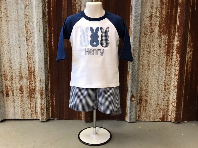 May include: A white and blue baseball style shirt with three blue bunny silhouettes and the name "Henry" embroidered on the front. The shirt is paired with blue and white gingham shorts.