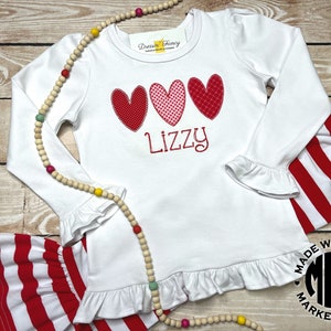 Puede incluir: Camiseta blanca de manga larga con corazones rojos y rosas y el nombre "Lizzy" bordado en la parte delantera. La camiseta tiene un dobladillo y mangas con volantes. La camiseta se combina con pantalones de rayas rojas y blancas.