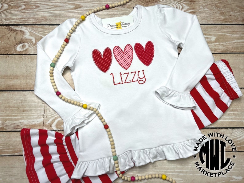 Puede incluir: Camiseta blanca de manga larga con corazones rojos y rosas y el nombre "Lizzy" bordado en la parte delantera. La camiseta tiene un dobladillo y mangas con volantes. La camiseta se combina con pantalones de rayas rojas y blancas.
