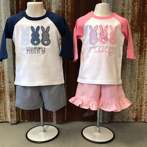 May include: Two children's outfits with a baseball tee shirt design featuring three bunny appliques. The boy's outfit is blue and white with the name "Henry" embroidered on the shirt. The girl's outfit is pink and white with the name "Lucy" embroidered on the shirt.