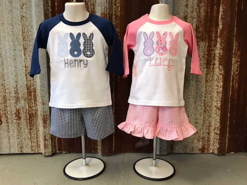 May include: Two children's outfits with a baseball tee shirt design featuring three bunny appliques. The boy's outfit is blue and white with the name "Henry" embroidered on the shirt. The girl's outfit is pink and white with the name "Lucy" embroidered on the shirt.