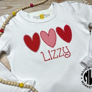 Puede incluir: Camiseta blanca de manga larga con tres corazones rojos y rosas y el nombre "Lizzy" bordado en la parte delantera. La camiseta tiene un pu&ntilde;o con volantes y un logotipo de "Made with Love Marketplace" en la parte inferior derecha.