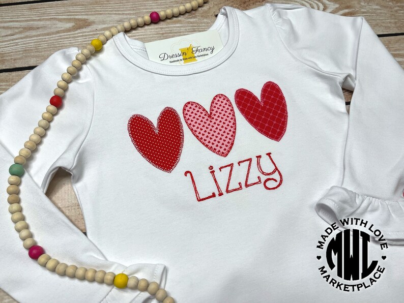 Puede incluir: Camiseta blanca de manga larga con tres corazones rojos y rosas y el nombre "Lizzy" bordado en la parte delantera. La camiseta tiene un pu&ntilde;o con volantes y un logotipo de "Made with Love Marketplace" en la parte inferior derecha.