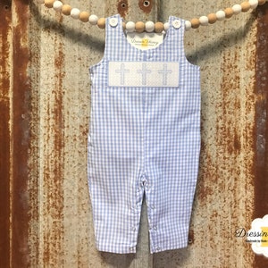 May include: A baby's blue and white gingham romper with three embroidered crosses on the chest. The romper has button closures on the straps and snaps at the legs. The background is a rustic, weathered metal.