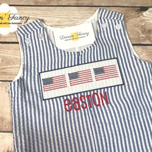 May include: A blue and white striped sleeveless romper with three embroidered American flags and the name "easton" embroidered below. The romper is made by Dressin' Fancy, a company that makes handmade items.