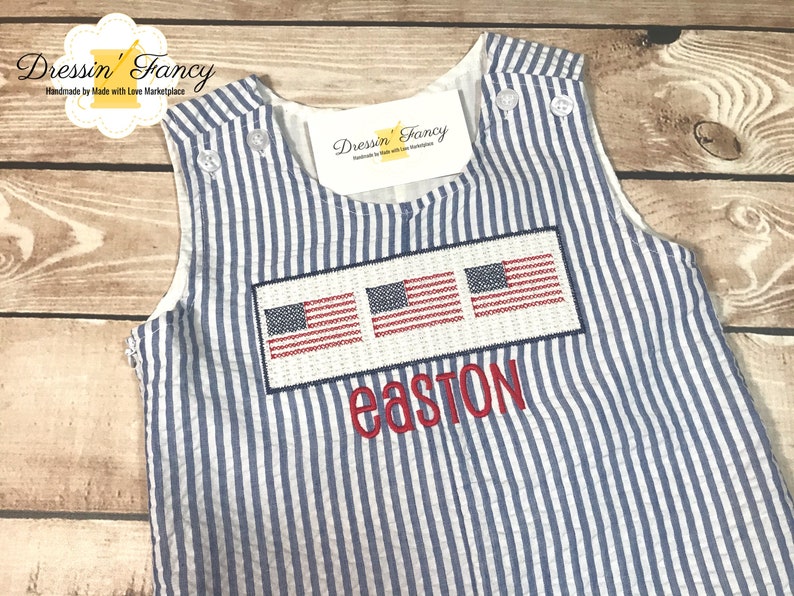 May include: A blue and white striped sleeveless romper with three embroidered American flags and the name "easton" embroidered below. The romper is made by Dressin' Fancy, a company that makes handmade items.