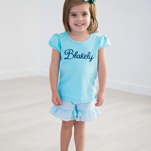 Puede incluir: Una ni&ntilde;a peque&ntilde;a con una camisa de manga corta azul claro con el nombre "Blakely" bordado en la parte delantera. Tambi&eacute;n lleva una falda de volantes a rayas azul claro y blanco. La ni&ntilde;a sonr&iacute;e y lleva un lazo azul claro en el pelo.