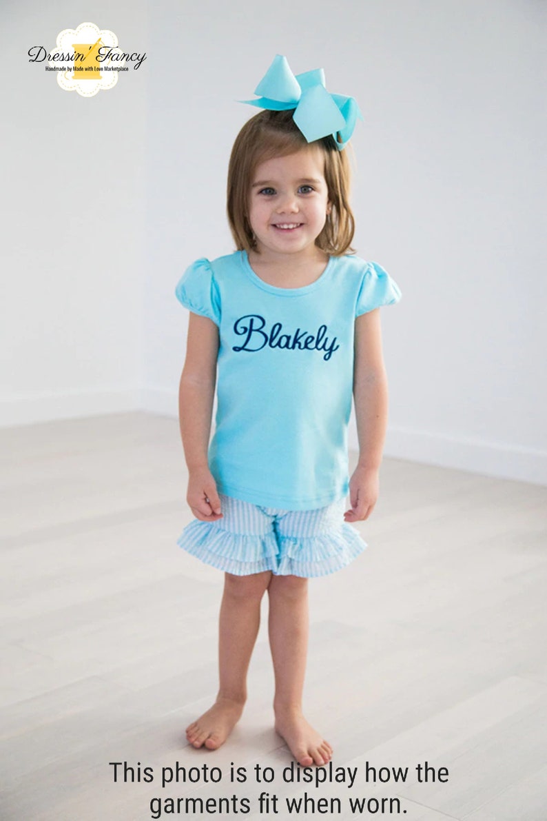 Puede incluir: Una ni&ntilde;a peque&ntilde;a con una camisa de manga corta azul claro con el nombre "Blakely" bordado en la parte delantera. Tambi&eacute;n lleva una falda de volantes a rayas azul claro y blanco. La ni&ntilde;a sonr&iacute;e y lleva un lazo azul claro en el pelo.