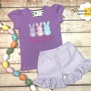 Puede incluir: Camiseta morada de manga corta con un gr&aacute;fico de tres peeps con forma de conejo de Pascua y el texto "Baby Blue". La camiseta se combina con pantalones cortos de rayas moradas y blancas con un dobladillo con volantes.