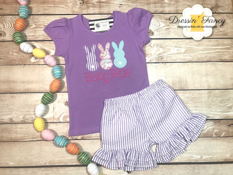 Puede incluir: Camiseta morada de manga corta con un gr&aacute;fico de tres peeps con forma de conejo de Pascua y el texto "Baby Blue". La camiseta se combina con pantalones cortos de rayas moradas y blancas con un dobladillo con volantes.