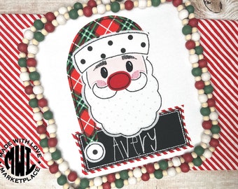 May include: A white Christmas-themed shirt featuring a Santa Claus face with a red nose and a plaid hat. The shirt includes a black name tag that says "Avery". The design is surrounded by a beaded garland and a red and white striped background.