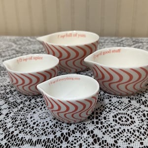 May include: Set of four white ceramic measuring cups with a wavy red pattern. Each cup has a different capacity marking and a whimsical phrase in red text: "1 cup full of yum", "1/3 cup of spice", "1/4 cup of everything nice", and "1/2 cup of good stuff".