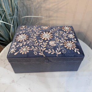 May include: A rectangular dark gray velvet jewelry box featuring intricate embroidery of floral patterns in shades of beige and brown, accented with small white beads at the center of each flower, and a small fabric tab on the front lid.