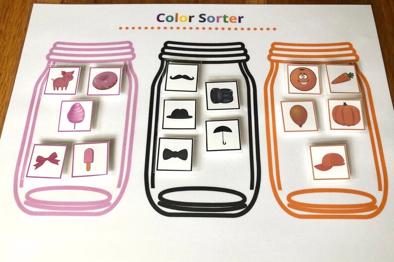 May include: A color sorting activity with three jars. The jars are labeled with a pink, black, and orange border. The jars contain various images of objects in different colors. The images include a pig, donut, cotton candy, bow, popsicle, mustache, hat, bow tie, umbrella, orange, carrot, balloon, pumpkin, and a baseball cap.