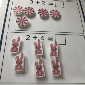 May include: A white board with two math problems. The first problem is 3 + 2 = with a picture of 5 red and white peppermint candies. The second problem is 2 + 4 = with a picture of 6 pink bunnies.