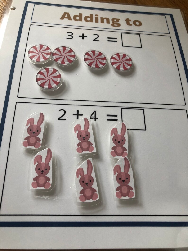 May include: A white board with two math problems. The first problem is 3 + 2 = with a picture of 5 red and white peppermint candies. The second problem is 2 + 4 = with a picture of 6 pink bunnies.