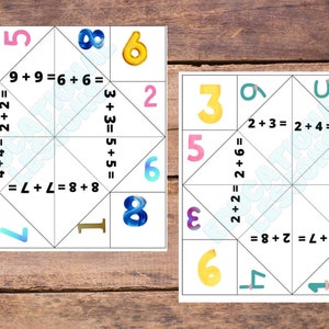 Può includere: Due indovinelli di carta bianca con numeri colorati ed equazioni matematiche. Gli indovinelli sono piegati e pronti per giocare. I numeri sugli indovinelli sono 2, 3, 4, 5, 6, 7, 8 e 9. Le equazioni sono semplici problemi di addizione.