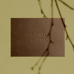 May include: A brown rectangular card with the embossed text "MATRON OF HONOR" is centered against a pale yellow background. Silhouetted branches are in the upper right corner.
