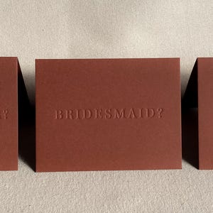 May include: Three brown cards with the text "Maid of Honor?", "Bridesmaid?", and "Matron of Honor?" embossed in white.