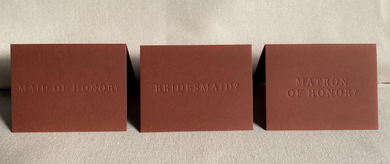 May include: Three brown cards with the text "Maid of Honor?", "Bridesmaid?", and "Matron of Honor?" embossed in white.