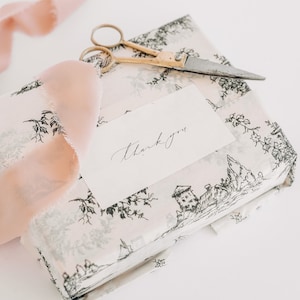 May include: A white gift box with a black and white floral pattern. The box is wrapped with a pink ribbon and a small card with the words "thank you" written on it. A pair of gold scissors and a small knife are also on top of the box.