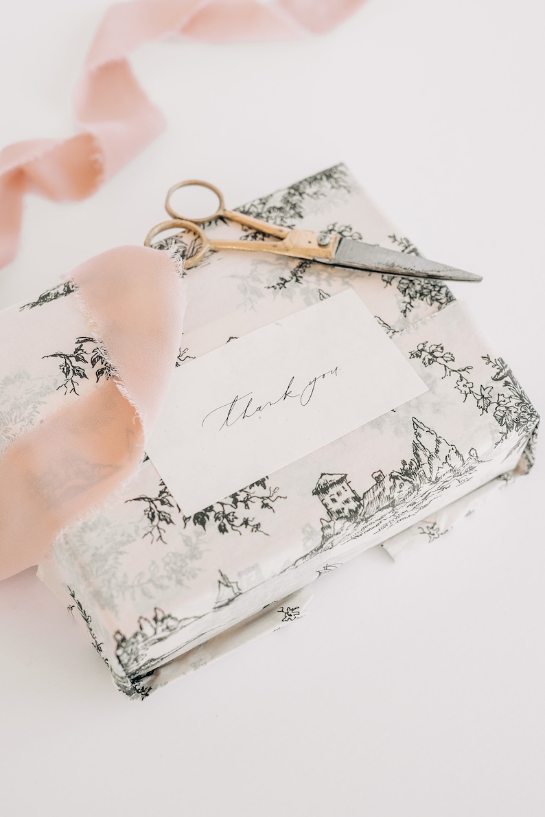 May include: A white gift box with a black and white floral pattern. The box is wrapped with a pink ribbon and a small card with the words "thank you" written on it. A pair of gold scissors and a small knife are also on top of the box.