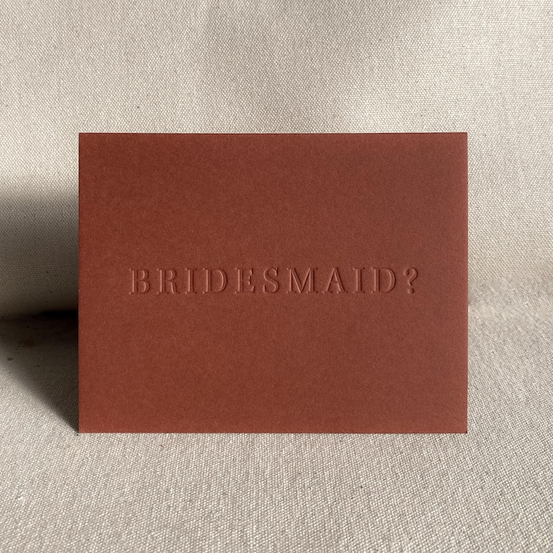 May include: A red card with the text "BRIDESMAID?" embossed in white.