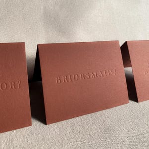 May include: Three folded cards with a brown textured surface. The text "BRIDESMAID?" is embossed on the center card.