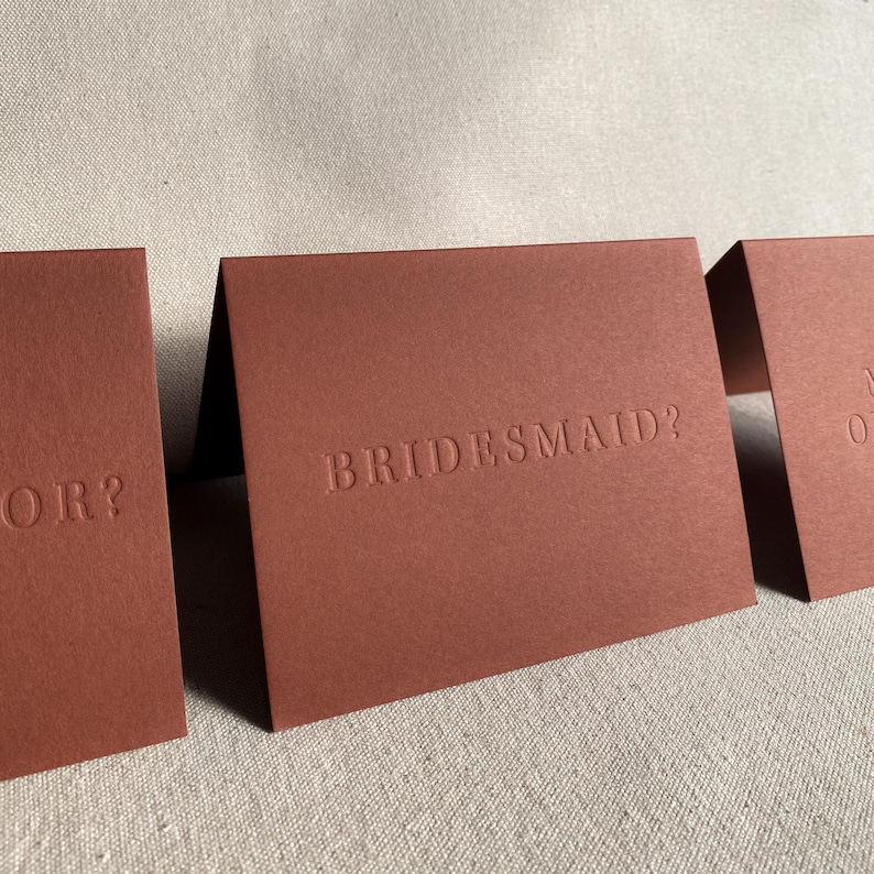 May include: Three folded cards with a brown textured surface. The text "BRIDESMAID?" is embossed on the center card.