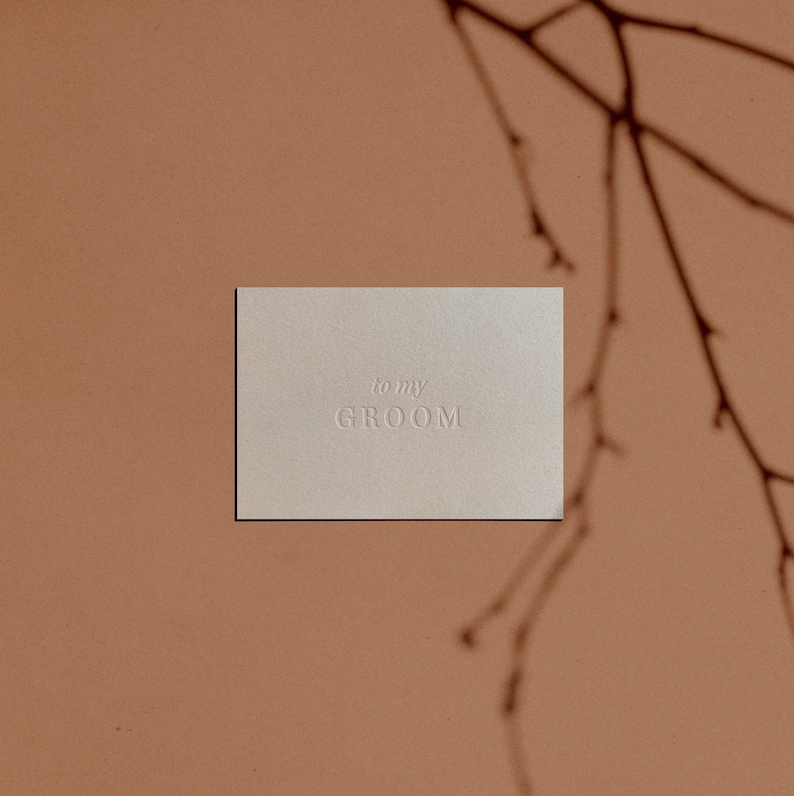 May include: A white card with the text "to my GROOM" embossed on it. The card is sitting on a light brown surface.