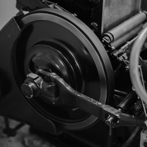 May include: A close-up of a large, black metal gear with a smaller gear attached to it. The gears are part of a printing press.